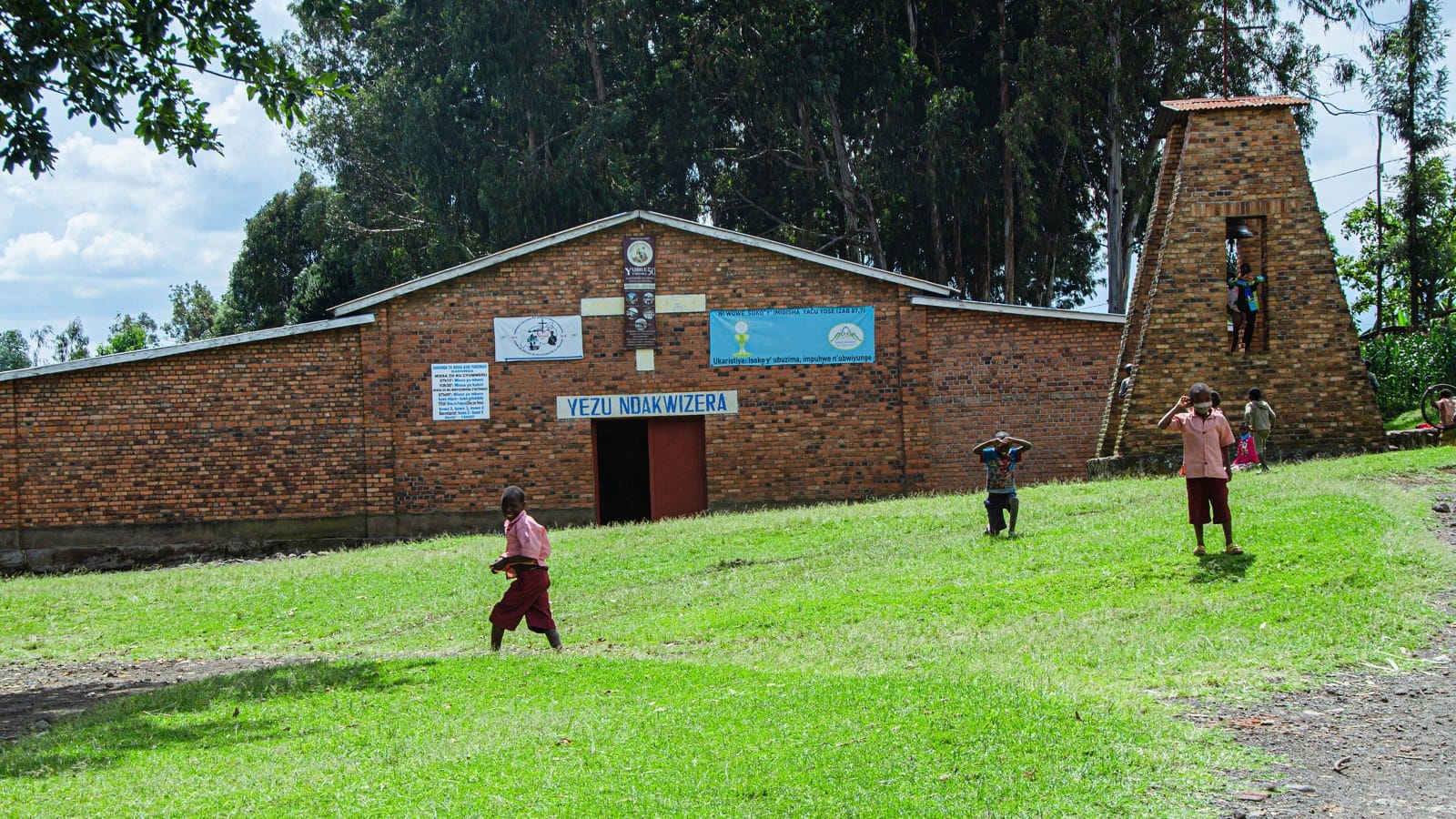 A rural brick church with a separate bell tower on a grassy hillside in Rwanda, with children playing in front
