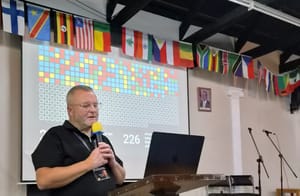 Kenno Leier speaking at a lectern in front of a slide of coloured blocks, with national flags along the wall